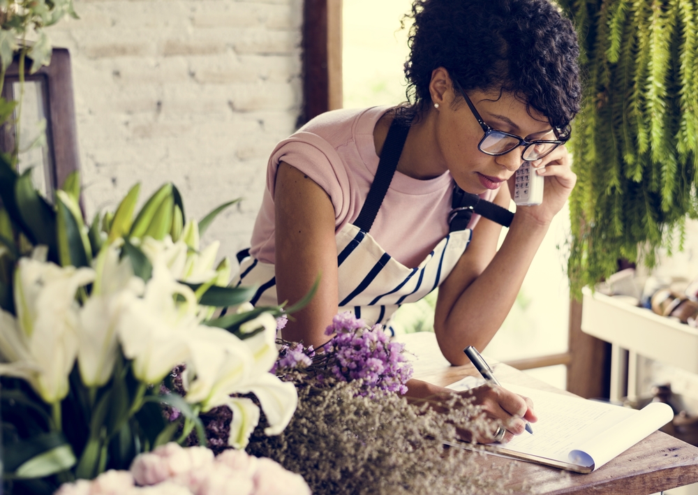 A women on the phone, writing information down on a notebook, surrounded by flowers