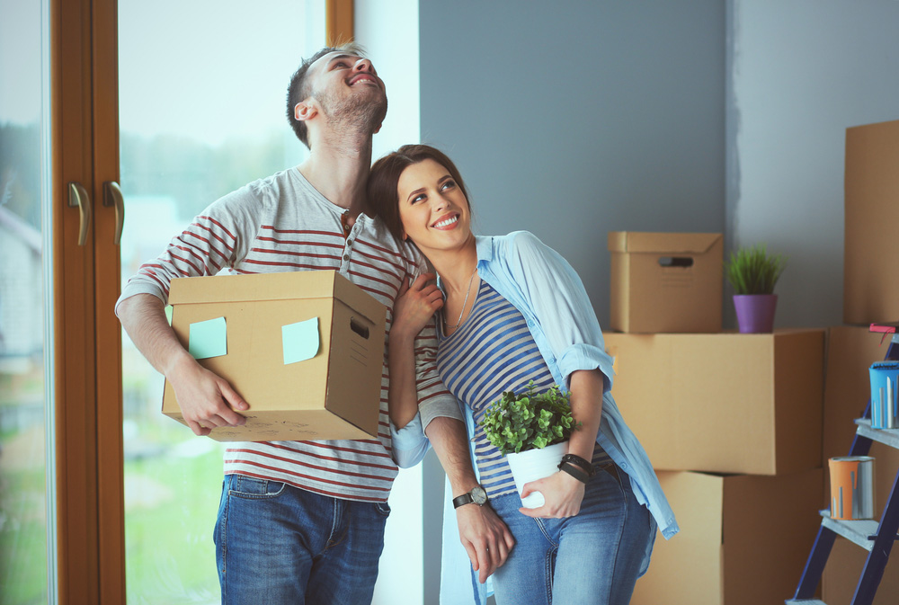 A man holding a box and a woman holding a plant smiling as they move into their new home
