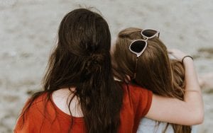 Two women sitting on a beach, facing away, resting on one another.