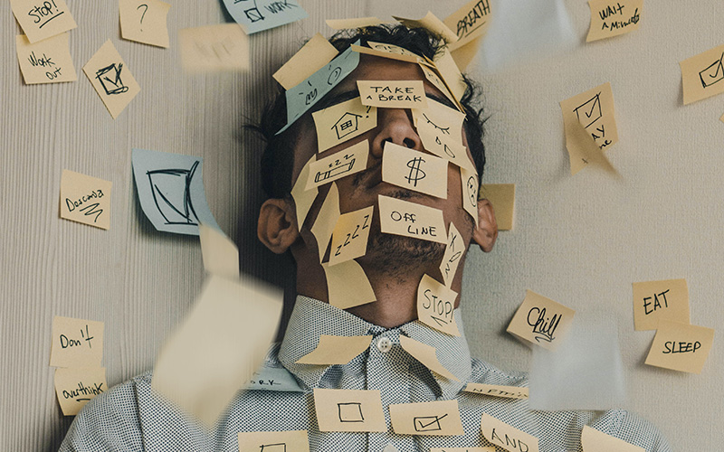 A bearded man laying down, overwhelmed by dozens of sticky-notes stuck to his face, each containing a responsibility.