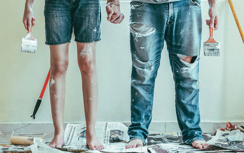 Two people holding hands and wearing paint-splashed jeans, with paintbrushes in hand.