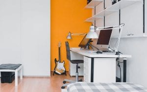 A tidy office, painted white, with a yellow feature wall. 2 Computers sit on the desk, and a guitar rests against the wall.