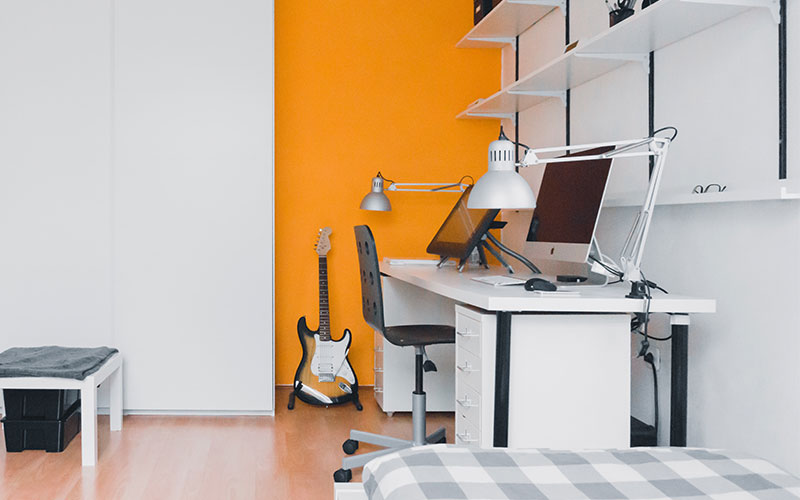 how-to-educate-your-children-on-the-cost-of-living-800x500 A tidy office, painted white, with a yellow feature wall. 2 Computers sit on the desk, and a guitar rests against the wall.