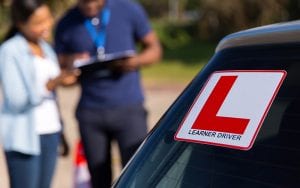 A car window with learner plates attached. Behind stands an instructor and a student having a conversation.