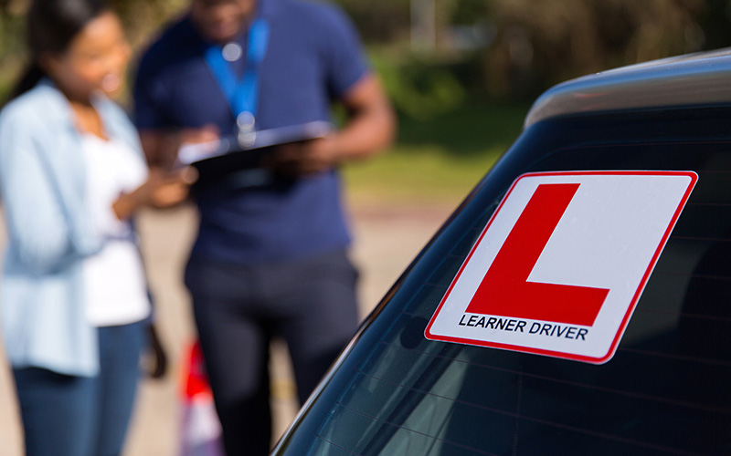 starting-your-journey-on-the-road-whilst-saving-cost-800x500 A car window with learner plates attached. Behind stands an instructor and a student having a conversation.