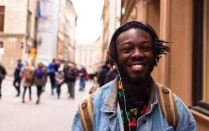 A man standing in public and smiling. He is wearing headphones and a backpack.