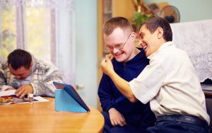 A father and son sit at a table while looking at a tablet, laughing together.
