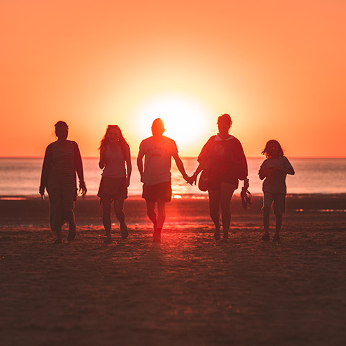 A group of five people walking towards the ocean, with the sun setting in front of them.