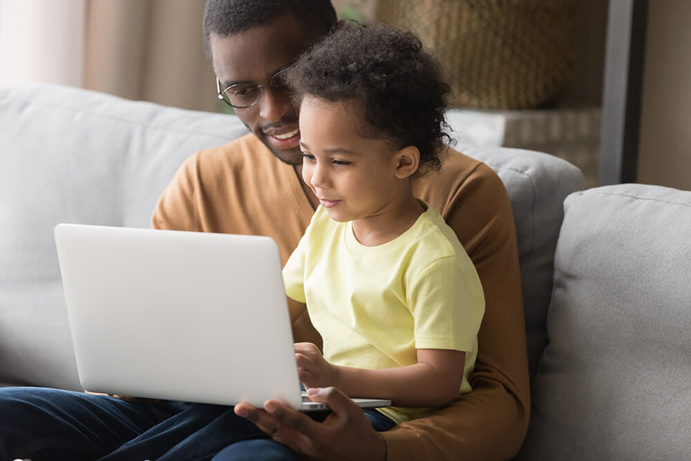 African father holds little son on lap family sitting on sofa at home with notebook, looking at screen watching cartoon educational videos, make video call, having fun enjoy free time together concept