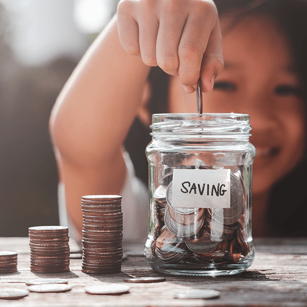 Young girl putting a coin in glass jar filled with money next to a stack of coins