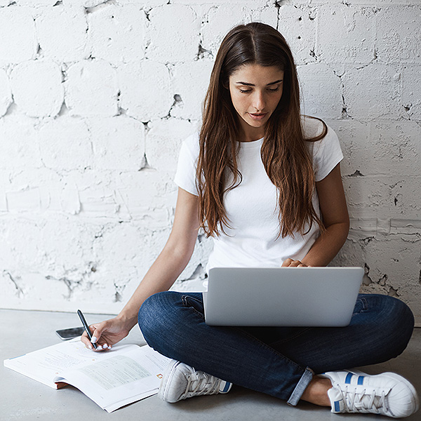 Woman sitting cross legged on the floor using laptop and writing on notepad