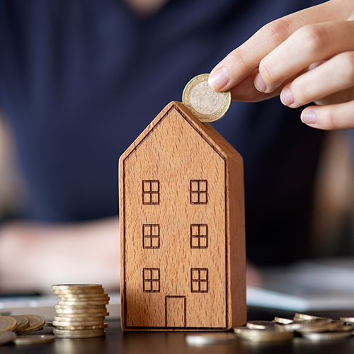 Hand putting coin in a wooden house money box next to a stack of coins