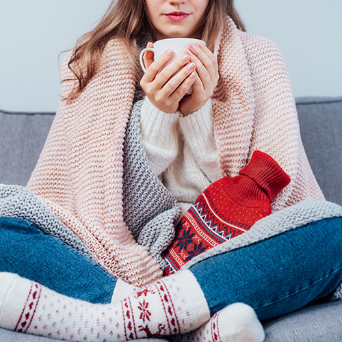 Woman freezes in wintertime. Young girl wearing warm woolen socks and wrapped into two blankets, holding a cup of hot drink and heating pad while sitting on sofa at home. Keep warm. Selective focus