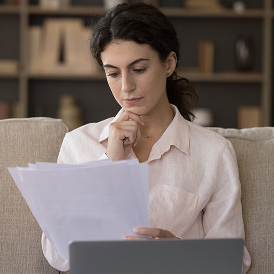 Serious focused young woman working with correspondence seated on sofa with wireless computer. Female holding papers, learn received formal document, analyze payment notification, do paperwork at home