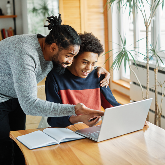Teenage boy on laptop. His father is leaning over him to look at the laptop, and has his arm around his son's shoulder.