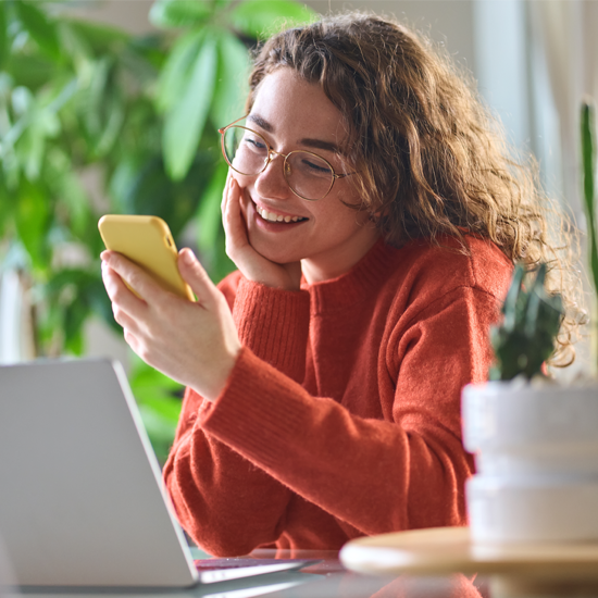 Young woman sitting in front of laptop. She's looking at her phone in her hand with a smile on her face