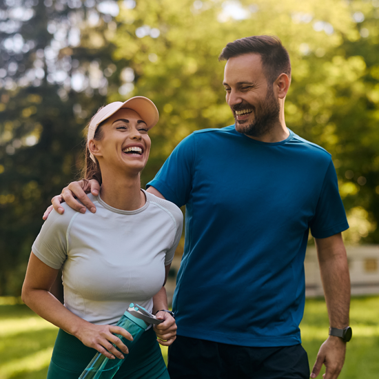 Man and woman dressed in sports clothing walking in the park. The man has his arm around the woman's shoulder and they are both laughing
