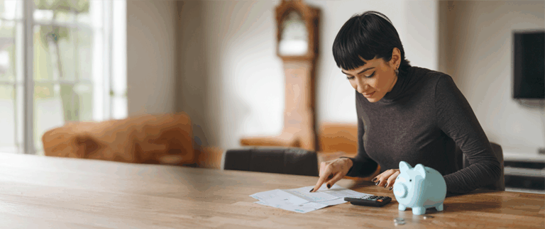 Person sitting at a wooden table reviewing finances with a calculator and a piggy bank