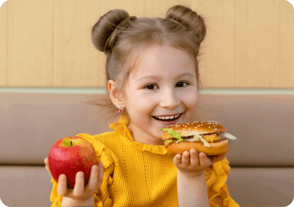 Child in a yellow top holding an apple in one hand and a burger in the other.