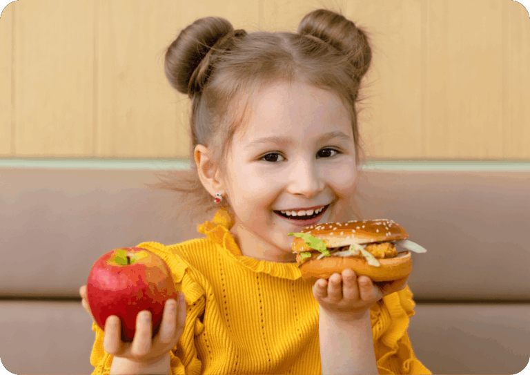 Child in a yellow top holding an apple in one hand and a burger in the other.