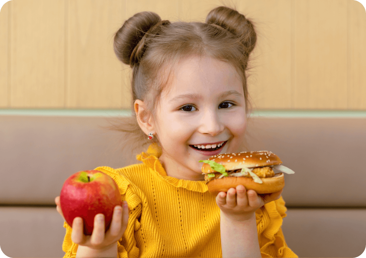 Child in a yellow top holding an apple in one hand and a burger in the other.