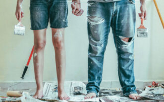 Two people holding hands and wearing paint-splashed jeans, with paintbrushes in hand.