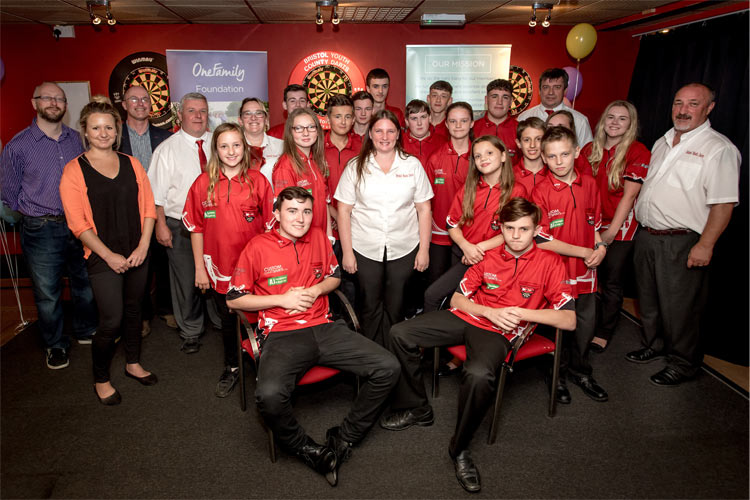 Dart club members sit in front of dart boards and OneFamily Foundation banners