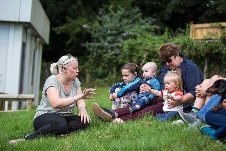 A woman sits on the grass teaching children sign language