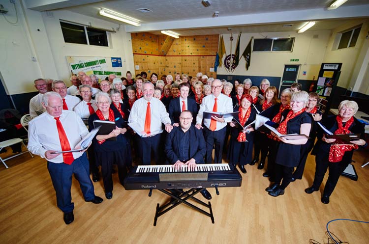 Members of Wythall Community Choir stand around a piano with their songbooks