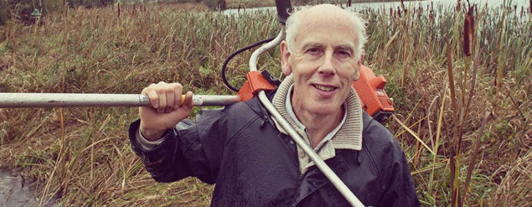 A man stands in front of some grass, carrying a gardening tool