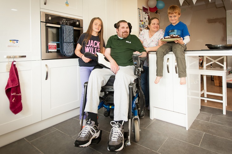 In a kitchen, two children and a woman stand with a man in a wheelchair