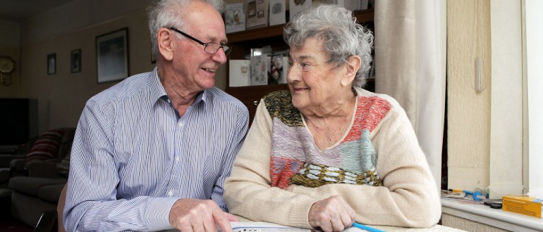 An elderly couple sit near to one another in their living room, near a window.
