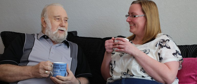 A bearded man and a women with glasses sit on a sofa enjoying a cup of tea