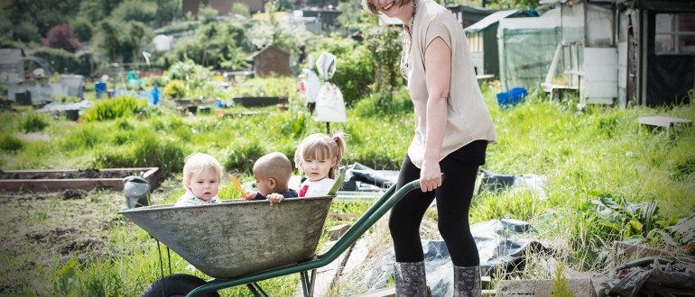 A lady on an allotment wheels three children along in a wheelbarrow.