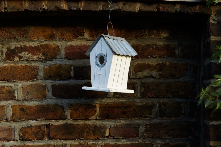 A bird feeder in the shape of a small house hangs in front of a brick wall.