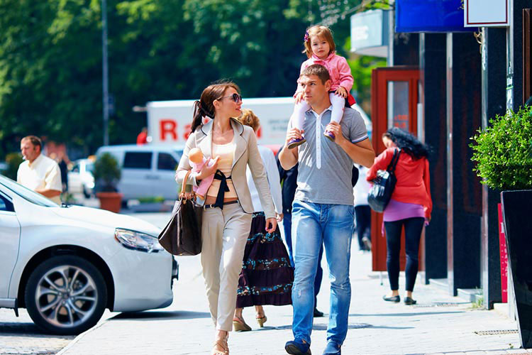 A young couple walk down the street. The man has their daughter on his shoulders.