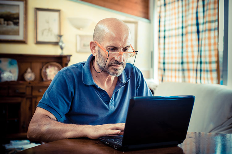 A man wearing spectacles sits at a table in his living room, looking at his laptop.