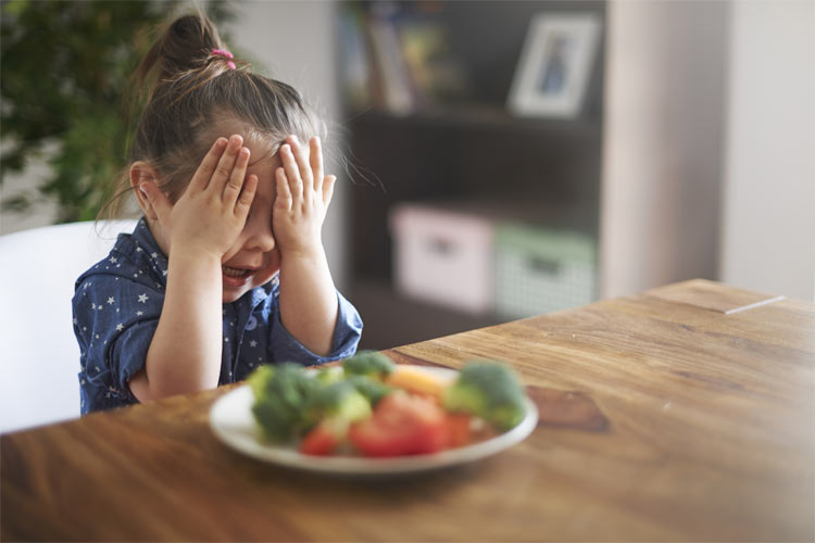 A little girl, presented with a plate of vegetables, hides her face.