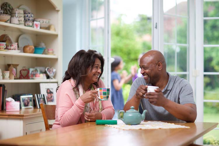 A couple having a cup of tea at the kitchen table, with patio doors open behind them.