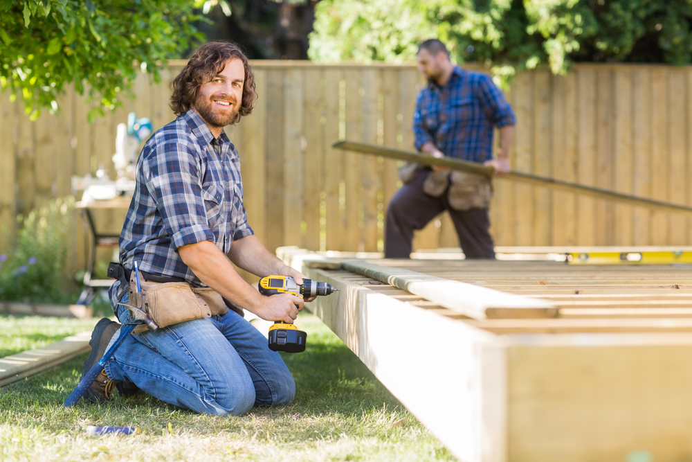 A bearded workman smiles at the camera while using his drill on a piece of decking. In the background, another workman carries a piece of wood across the garden.
