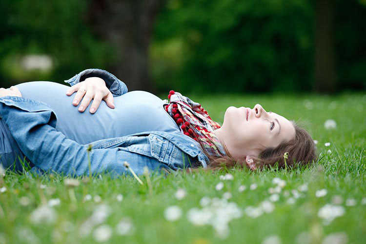 A pregnant woman lies on the grass, holding her belly and looking at the sky.