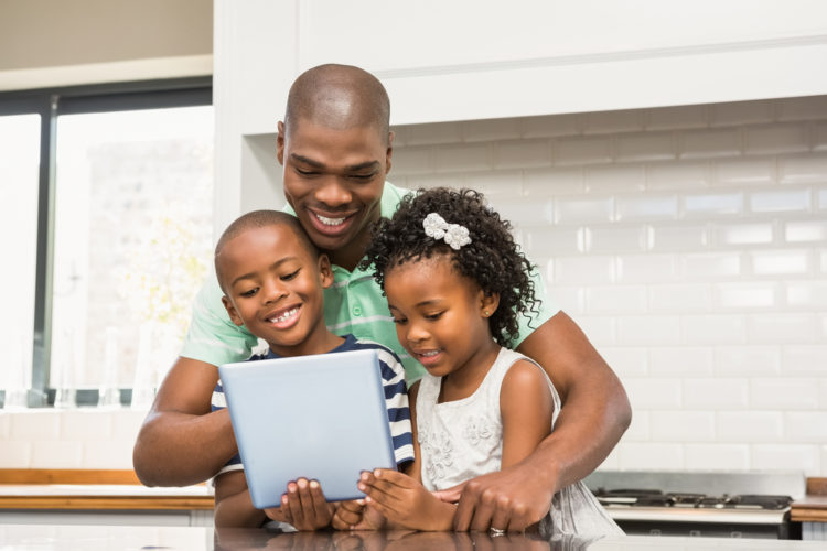 A man stands in his kitchen with his two children, looking at an iPad.