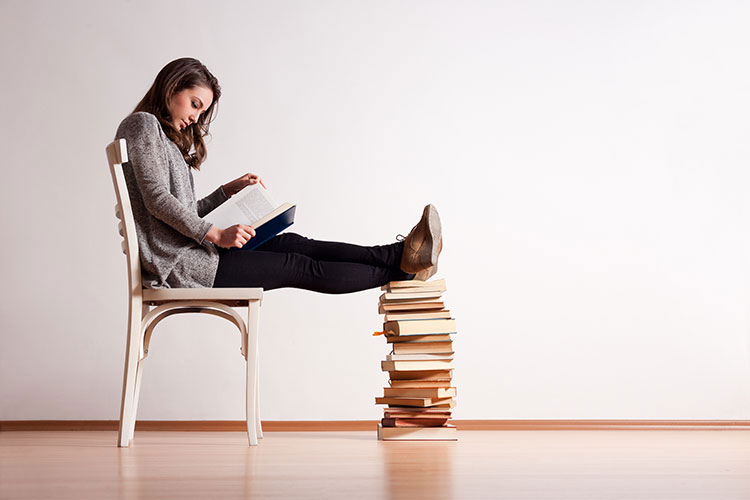 A student sitting on a chair, reading, with her feet up on a pile of books.