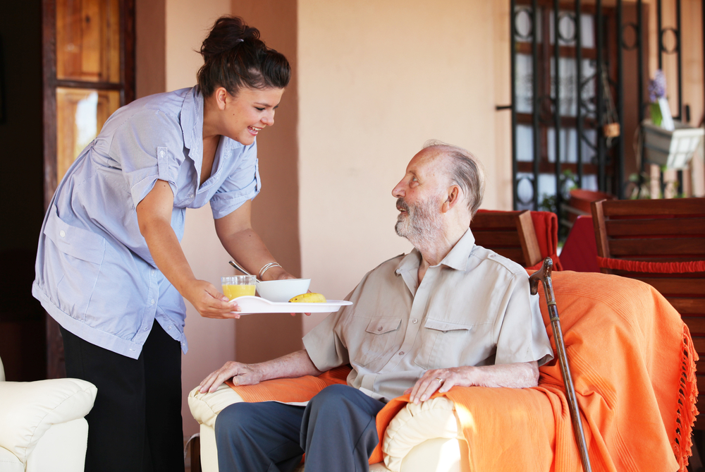 A nurse brings cereal and orange juice on a tray to an older man sitting on an armchair, his walking stick propped against the chair.