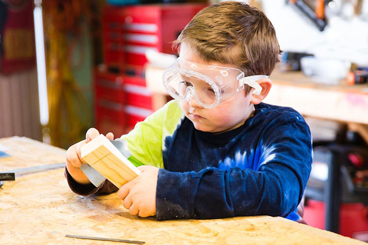 A little boy in goggles sanding a piece of wood.