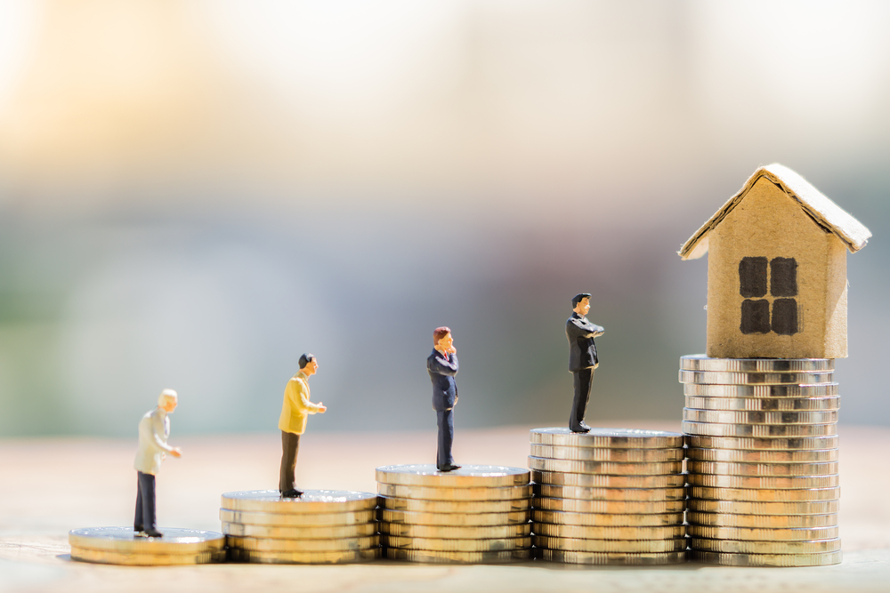Little figures stand on stacks of coins like steps leading up to a toy house.