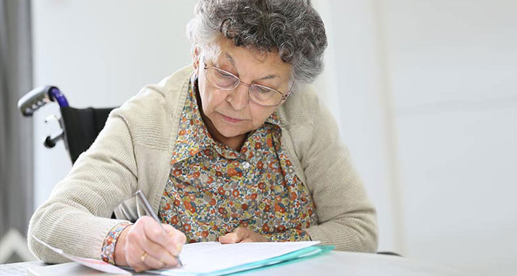 An older woman sits at a table filling out a form.
