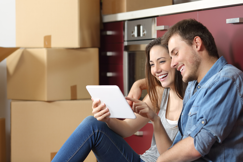 A young couple sit on the floor of their apartment, cardboard boxes in the background, smiling at something on the woman's iPad.