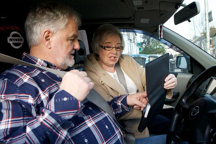 An older man in a car, putting on his seatbelt, while the woman in the passenger seat shows him something in a leather wallet.