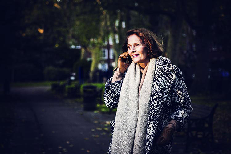 An older woman in a winter coat, talking on her mobile phone while standing the driveway of a large house.
